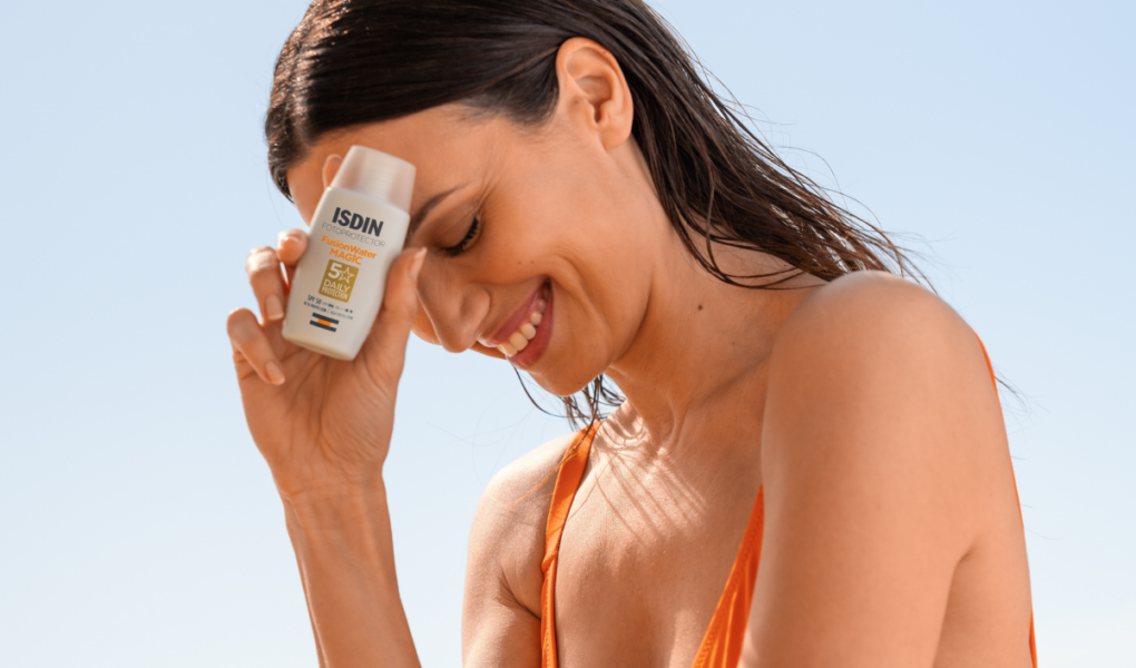 Mujer sujetando un producto de isdin, en la playa con el bikini puesto